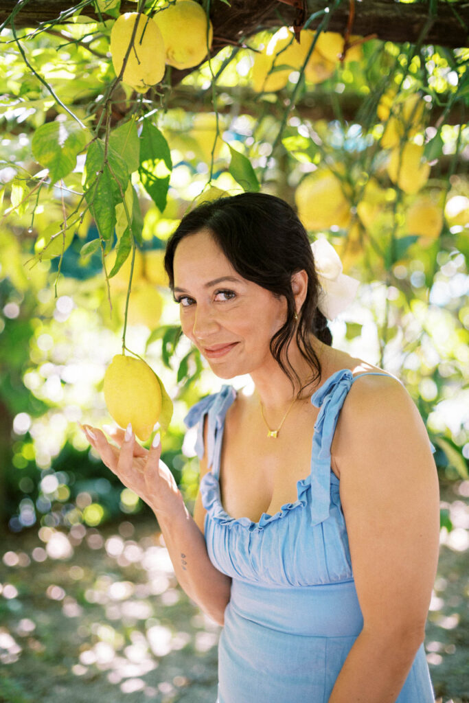 A woman in a blue dress in front of a lemon tree, holding a lemon in her hand.