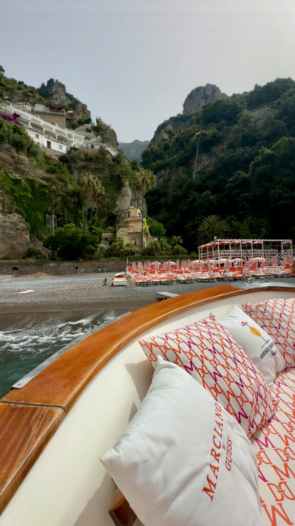 The side of a boat with pillows on the water in front of the cliffs and shore of the Amalfi Coast.