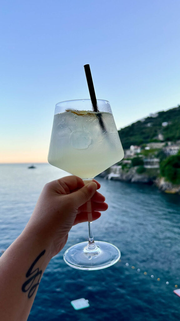 A hand holding a lemon cocktail in front of the ocean of the Amalfi Coast.