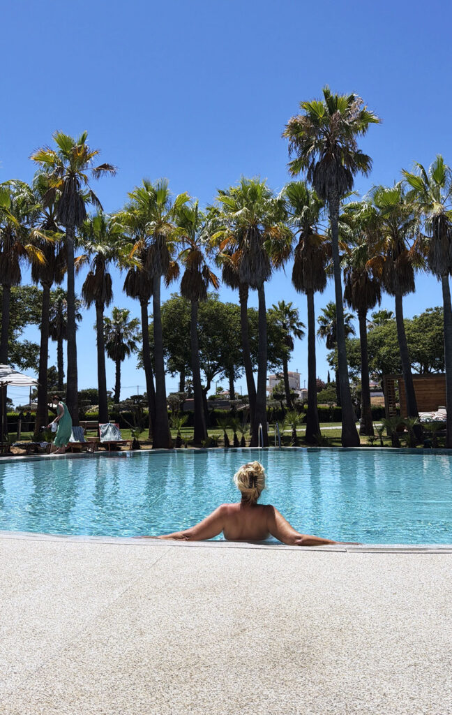 Woman relaxing in the pool with her back to the camera, palm trees and blue sky behind her, at Kimpton Atlântico Algarve