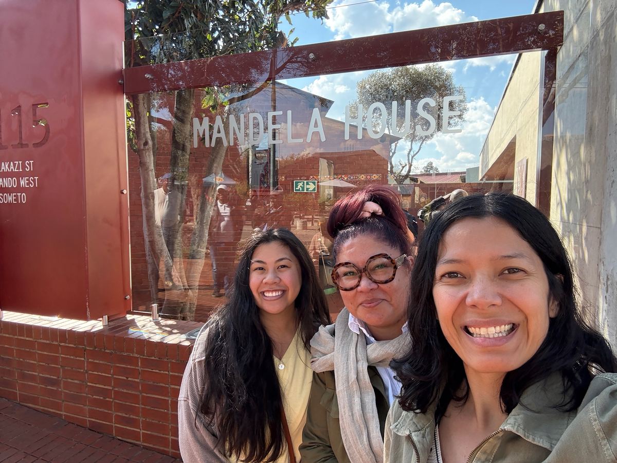 Three women smiling in front of the Mandela House sign in Soweto