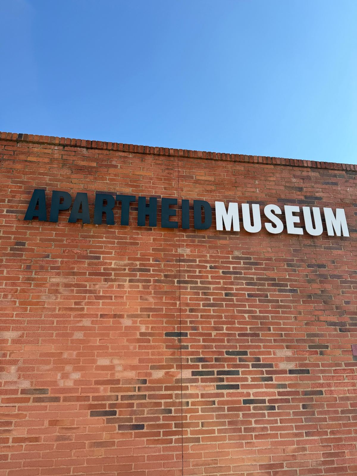 Apartheid Museum sign on a red brick wall under a blue sky