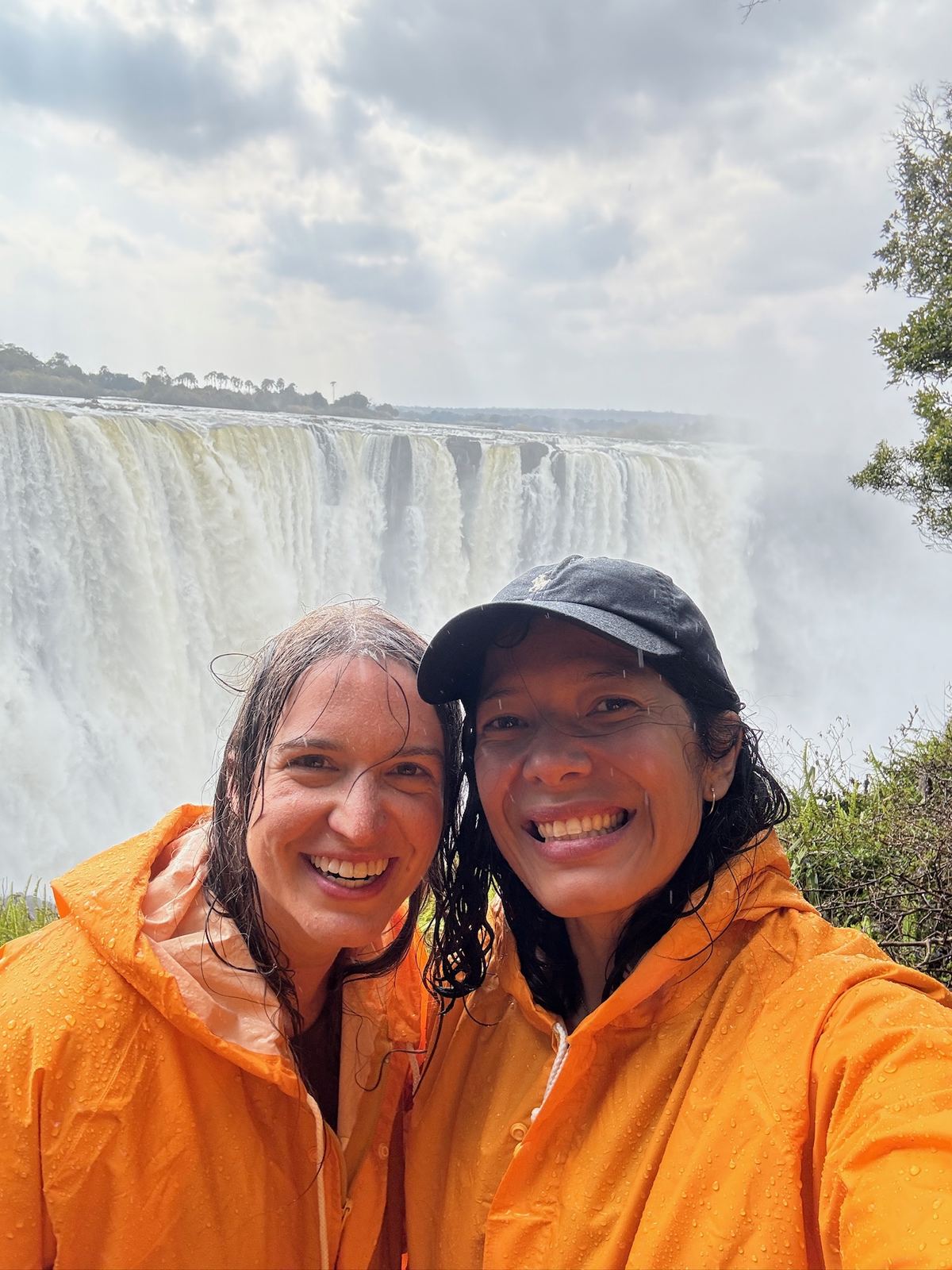 Two women in orange ponchos, soaked from the mist, smiling in front of Victoria Falls