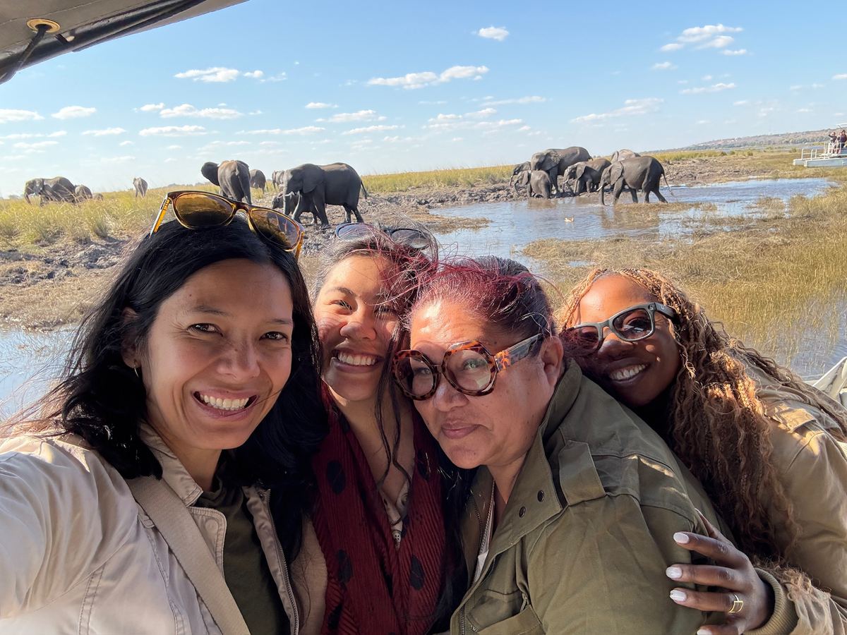 Four women taking a selfie on a boat with a herd of elephants on the Chobe River behind them