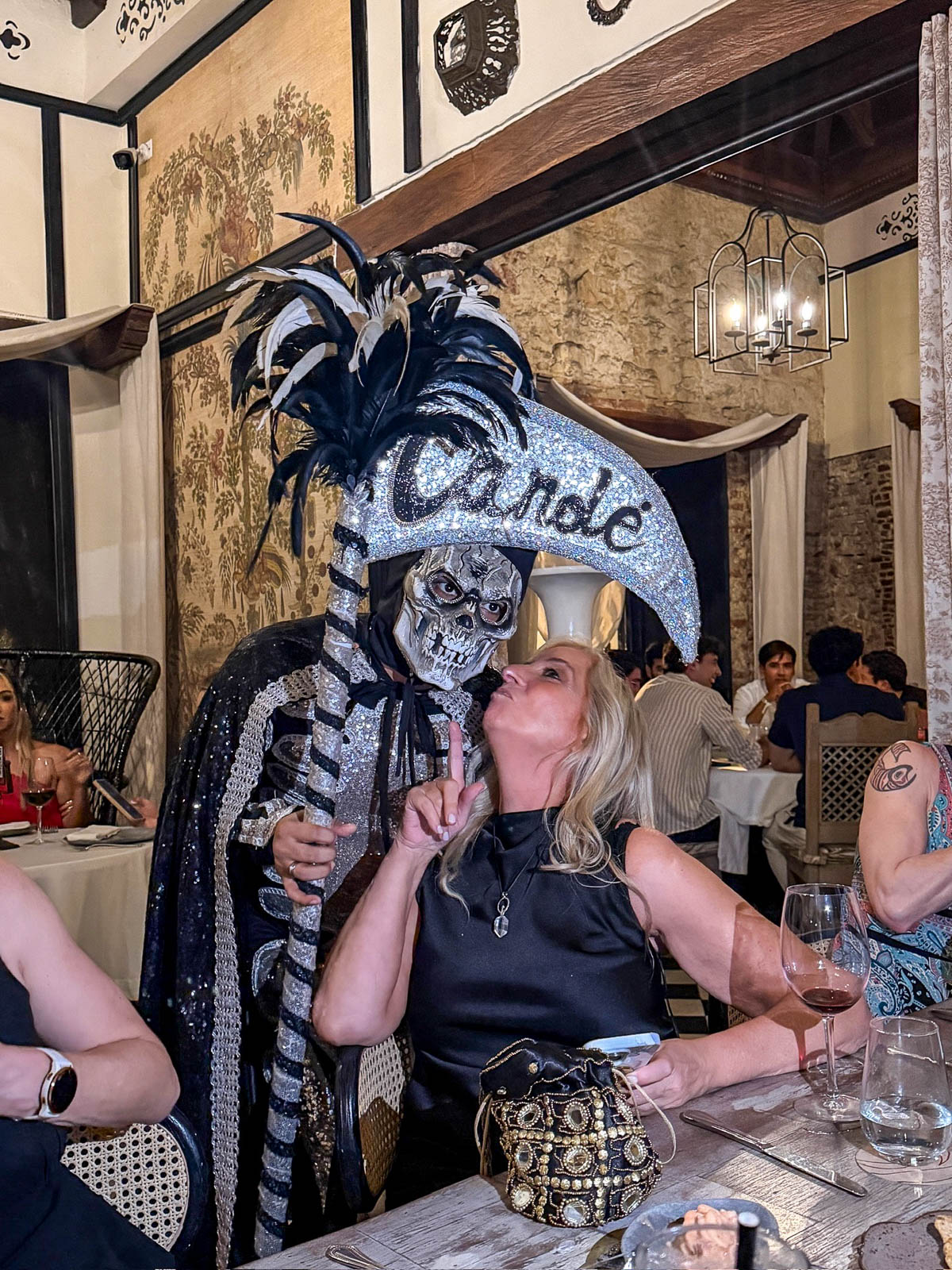 Masked performer in a feathered costume holding a Candé sign next to a smiling diner