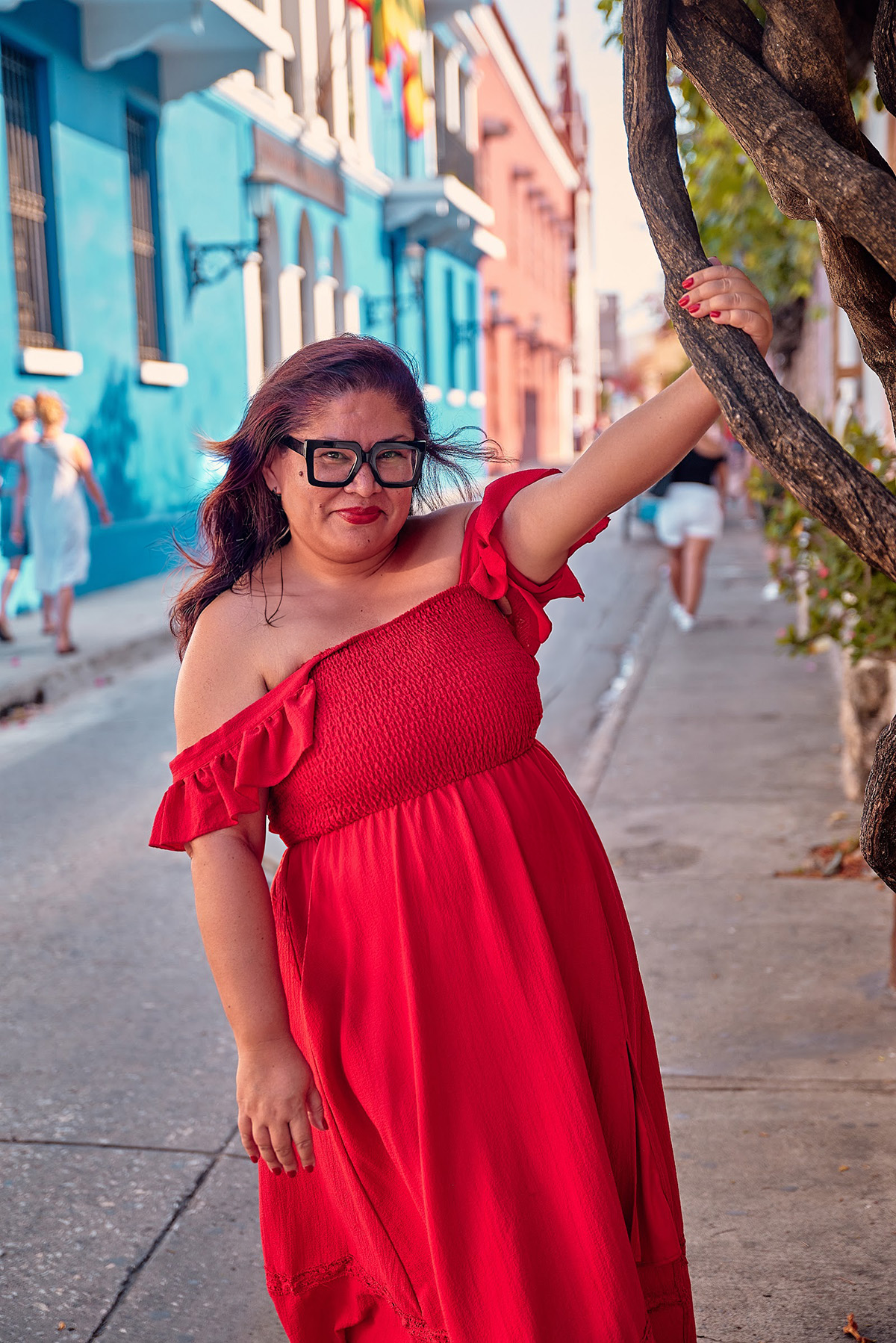 Cynthia in a red off the shoulder dress leaning on a twisted tree on a colorful street in Cartagena