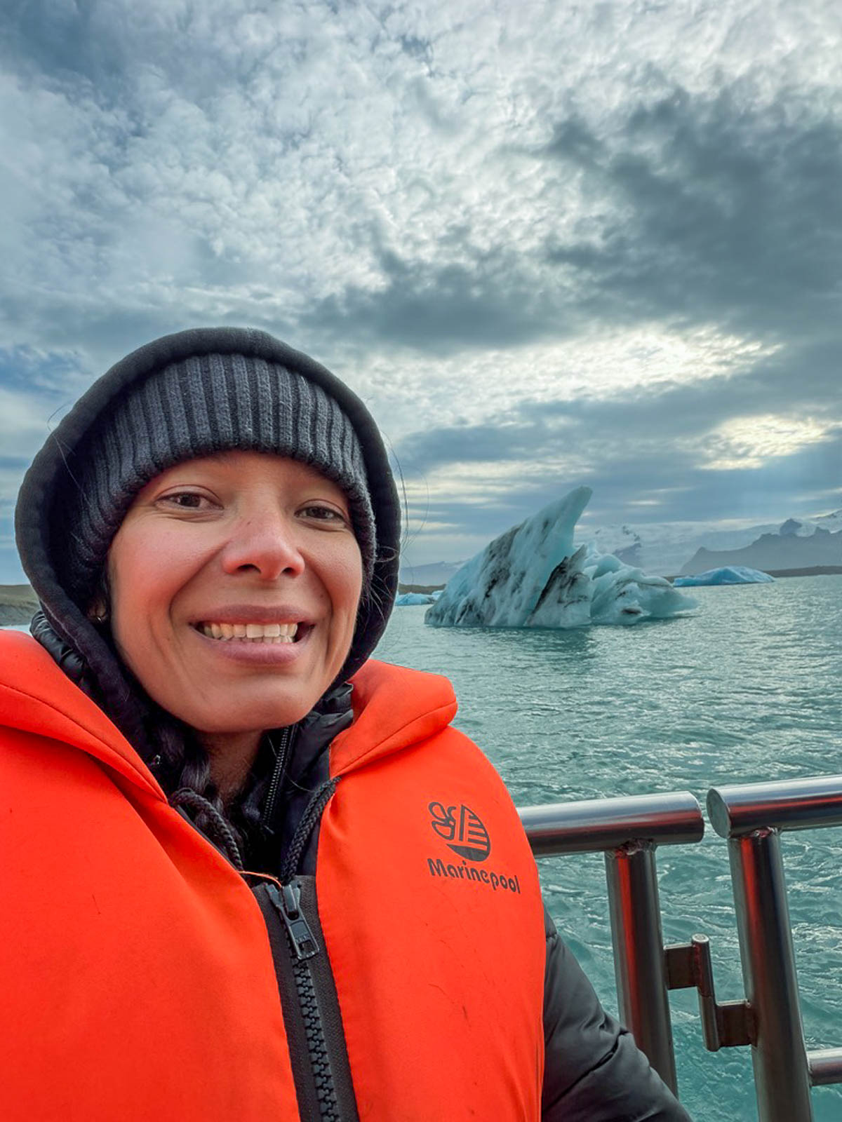 Toni in an orange flotation suit on the boat at Jökulsárlón Glacier Lagoon, with an iceberg behind her