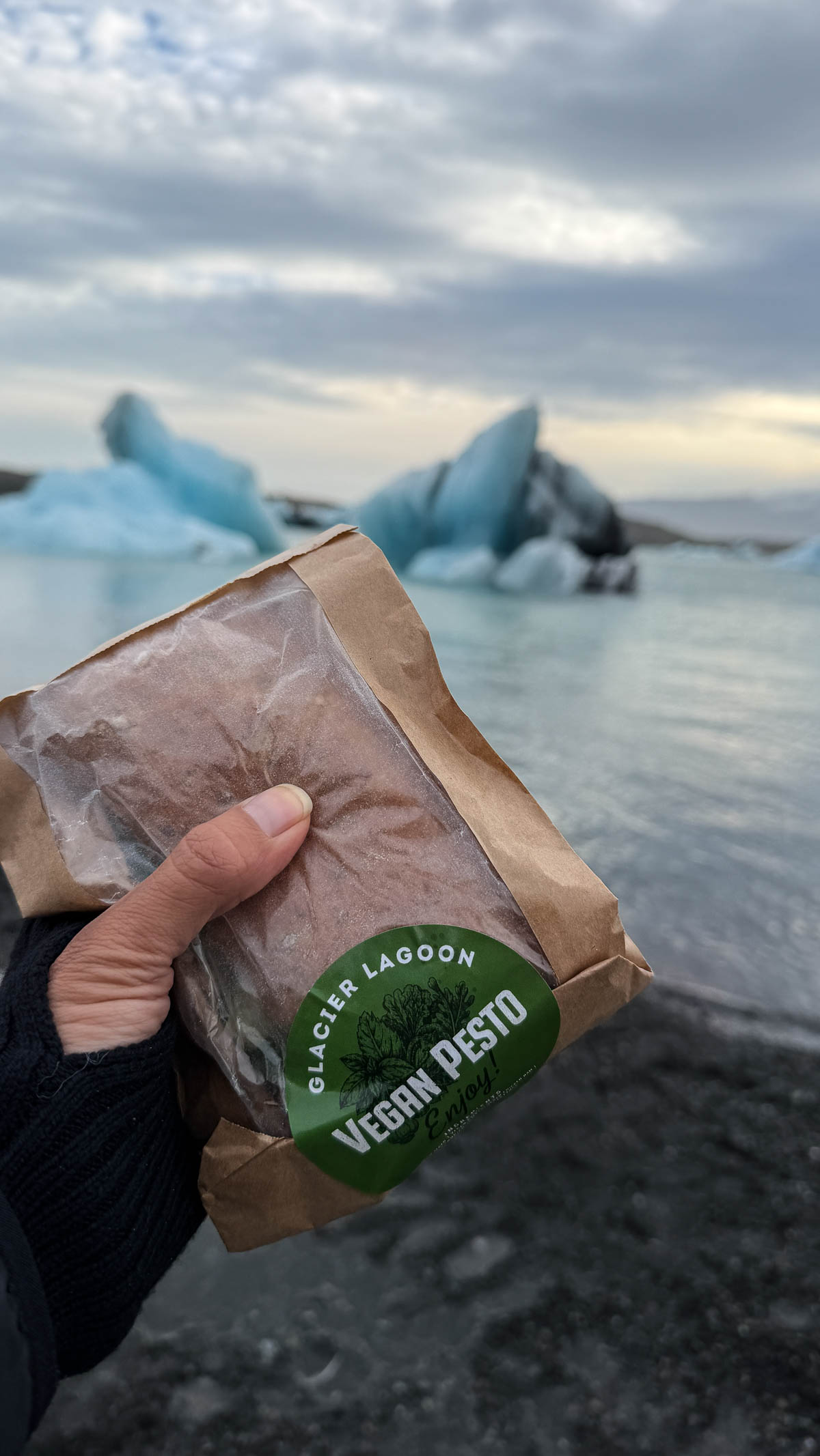 A hand holding a wrapped vegan pesto sandwich in front of the icebergs at Jökulsárlón Glacier Lagoon