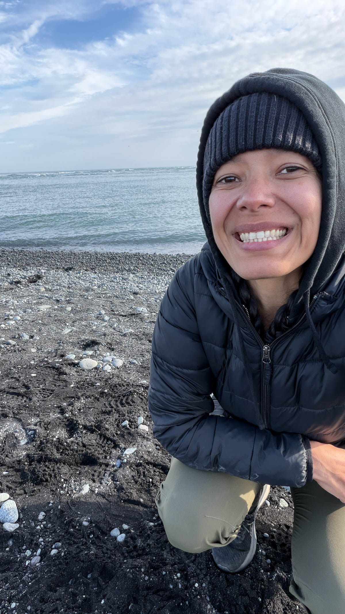 Toni crouching on a black-sand beach near the glaciers in Iceland with the ocean behind her