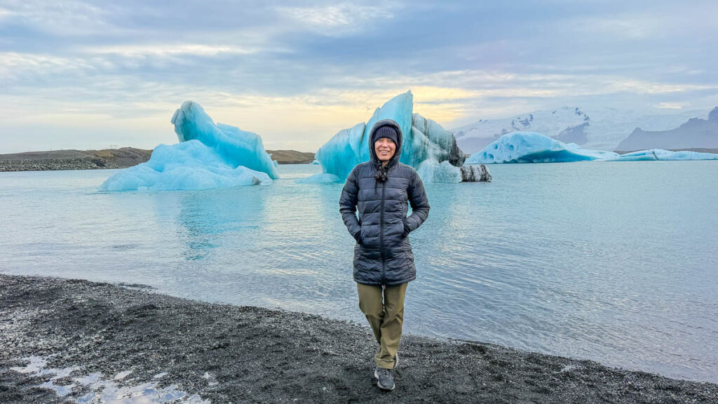 Toni standing on the black shore of Jökulsárlón Glacier Lagoon in Iceland with blue icebergs behind her
