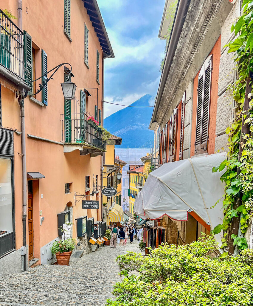 A sloping cobblestone street in Bellagio lined with peach buildings and shops, with Lake Como and the mountains at the bottom.