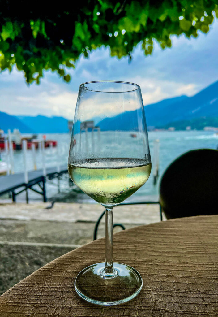 A glass of white wine on a table at the Bellagio waterfront, with boats and Lake Como behind it.