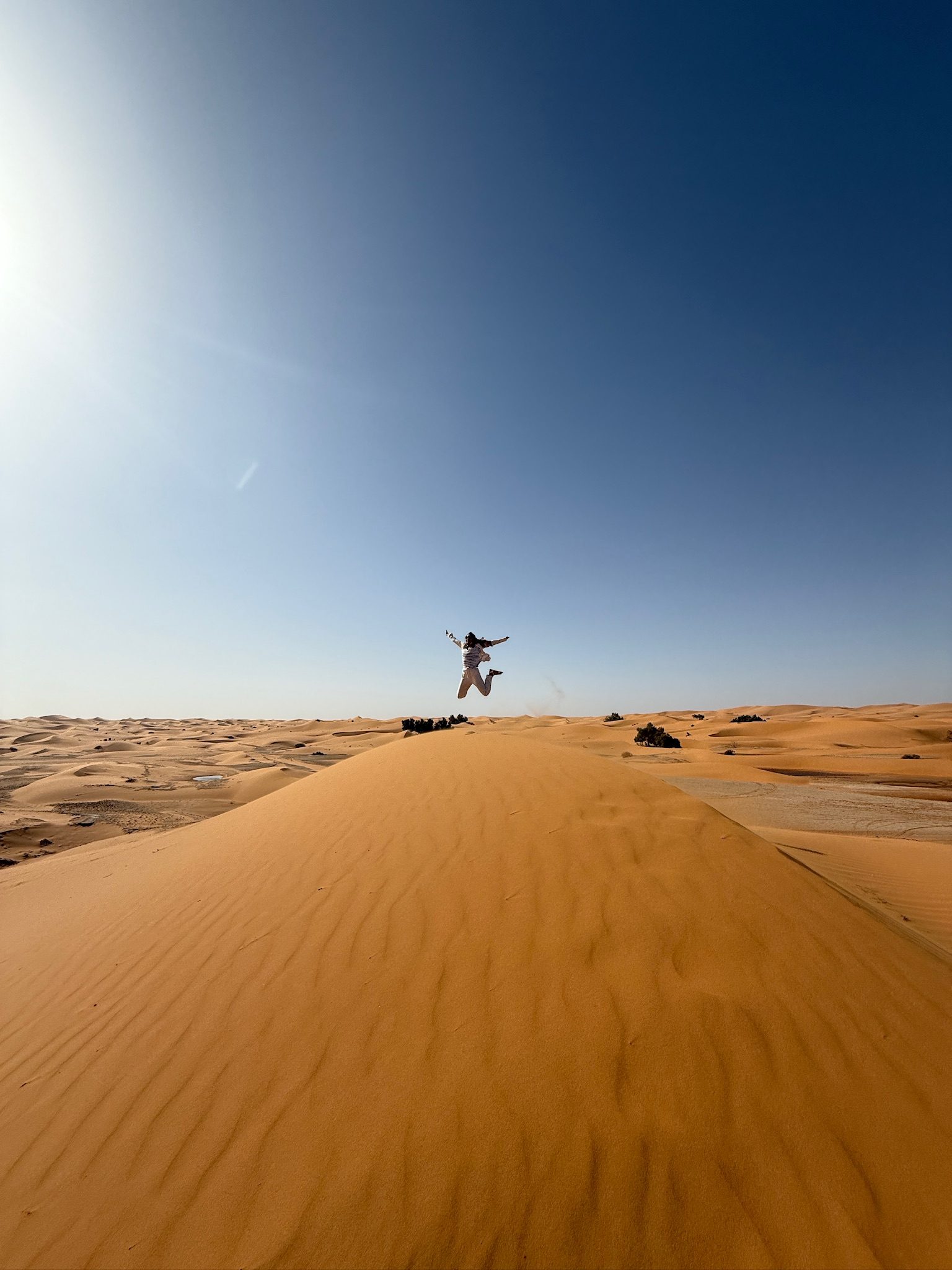 Person jumping on top of a tall sand dune under a clear blue sky