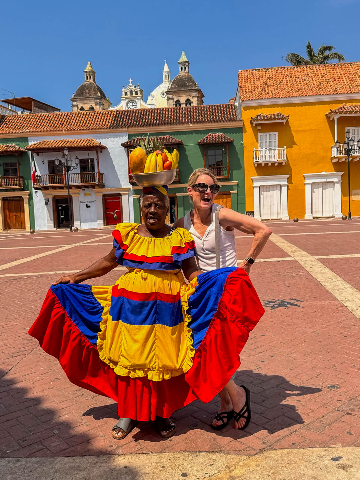 A palenquera spreading her bright yellow, blue, and red skirt next to a traveler in a plaza with the cathedral domes behind them in Cartagena