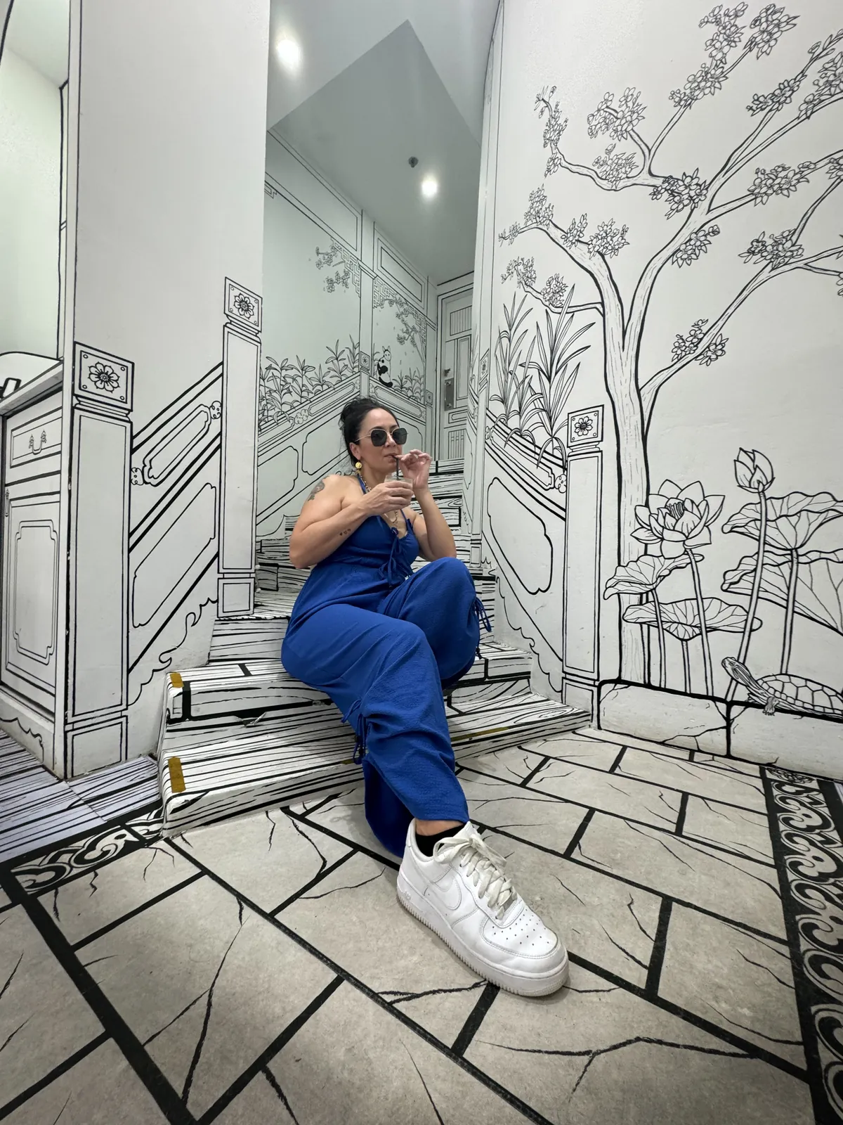 Liz in a blue jumpsuit sipping a drink on the hand drawn black and white stairs at Cafe Monochrome in Singapore