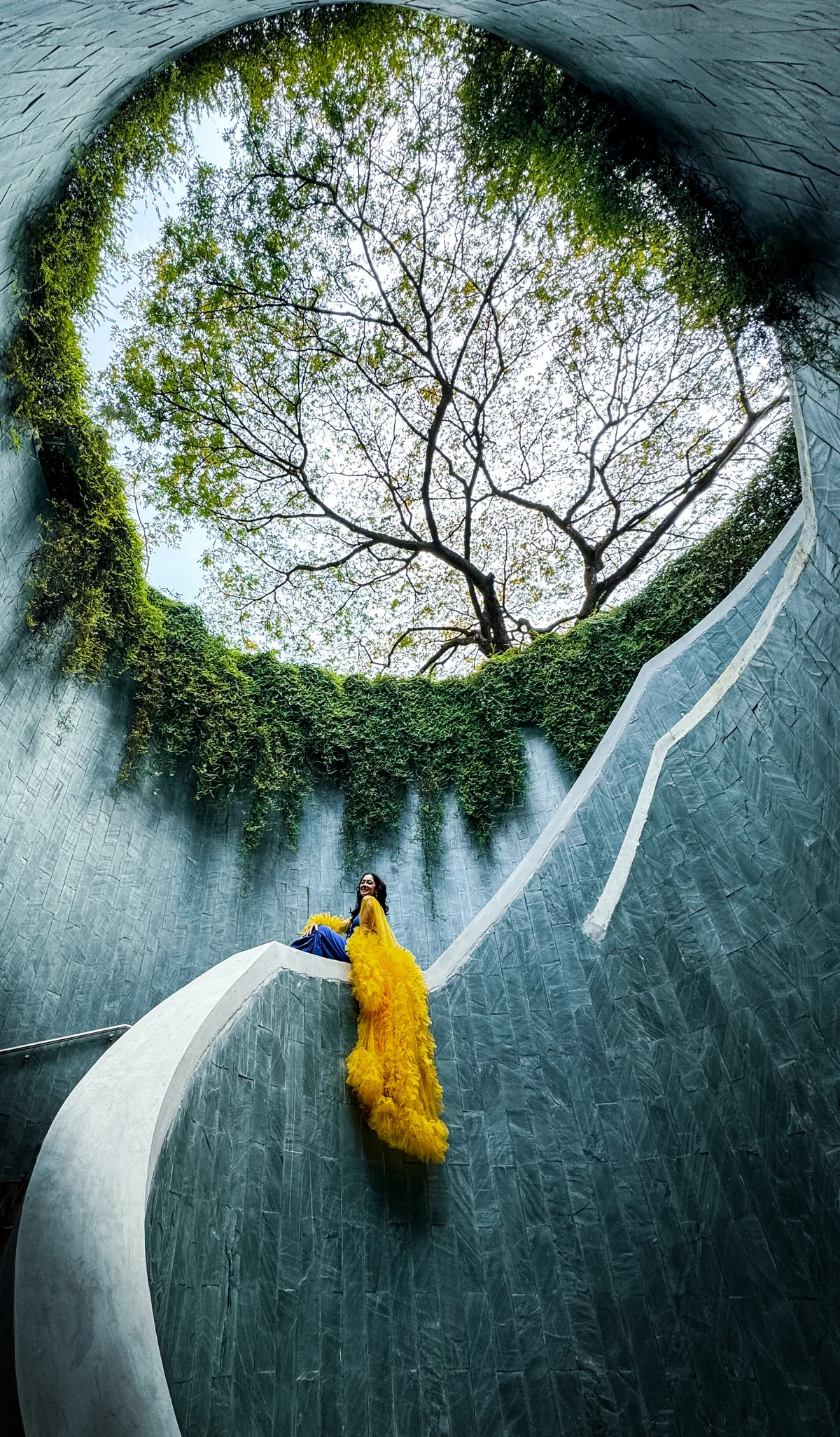 Liz in a yellow tulle dress on the spiral stair wall of the Fort Canning Park Tree Tunnel, looking up at the trees