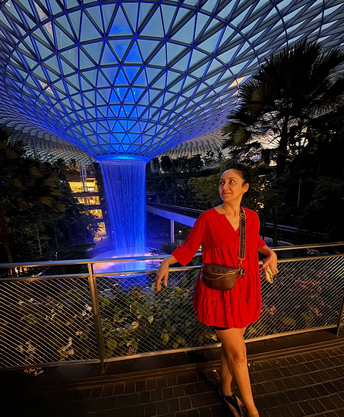 Liz in a red dress at night in front of the blue lit Rain Vortex at Jewel Changi Airport