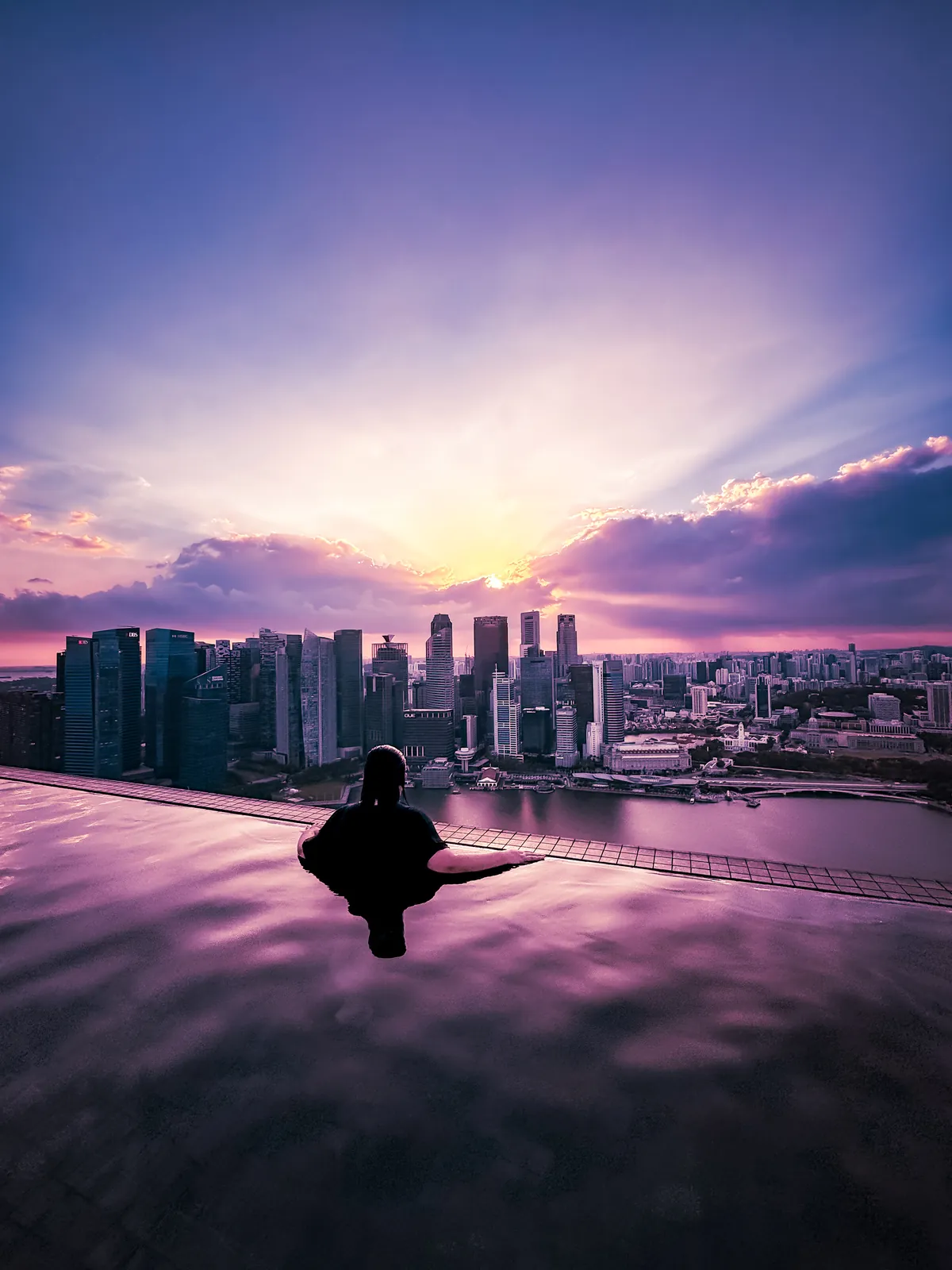 Cynthia silhouetted in the Marina Bay Sands Infinity Pool under a pink and purple sunset over the city skyline