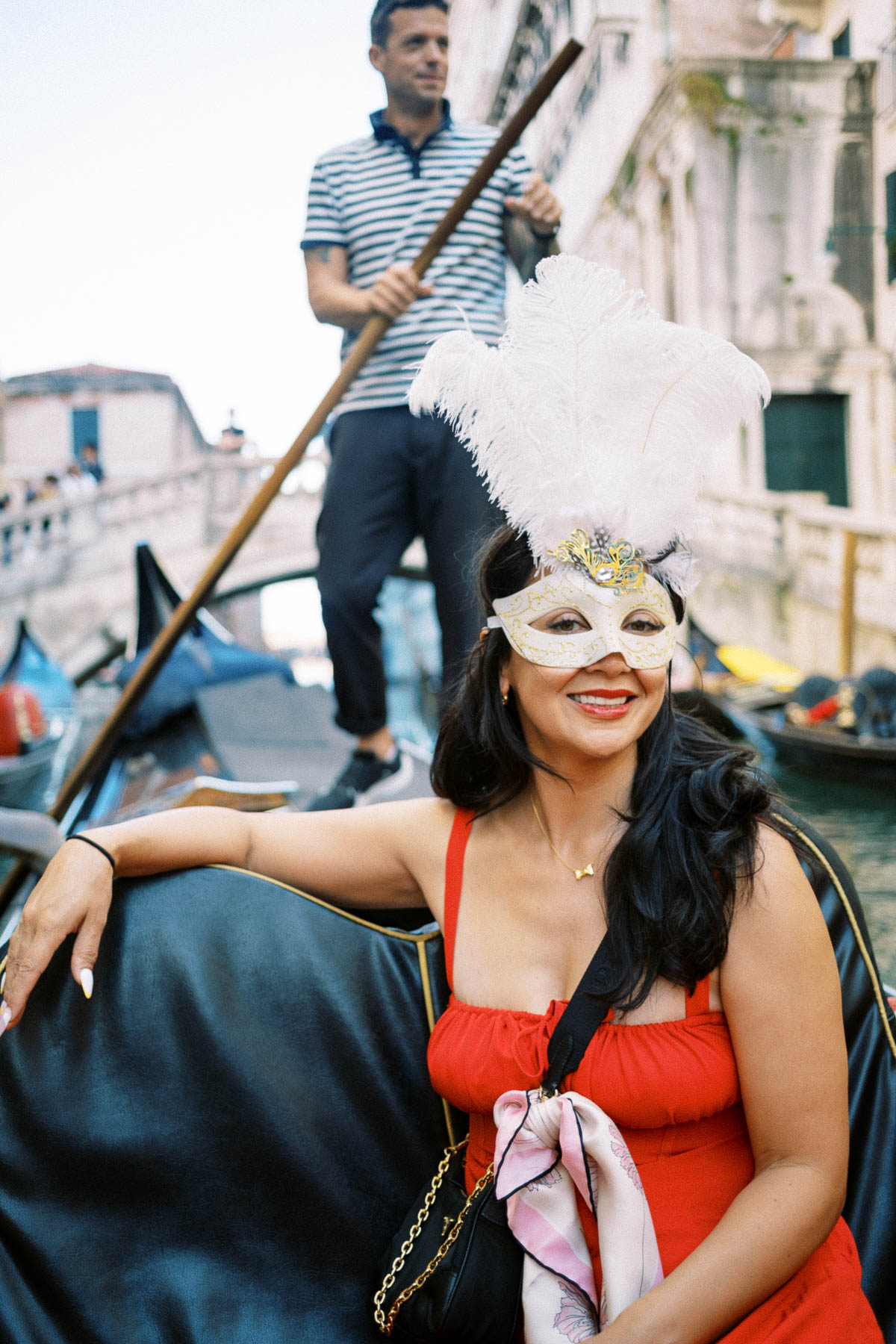 A traveler in a white feathered Venetian mask on a gondola with the gondolier rowing behind her