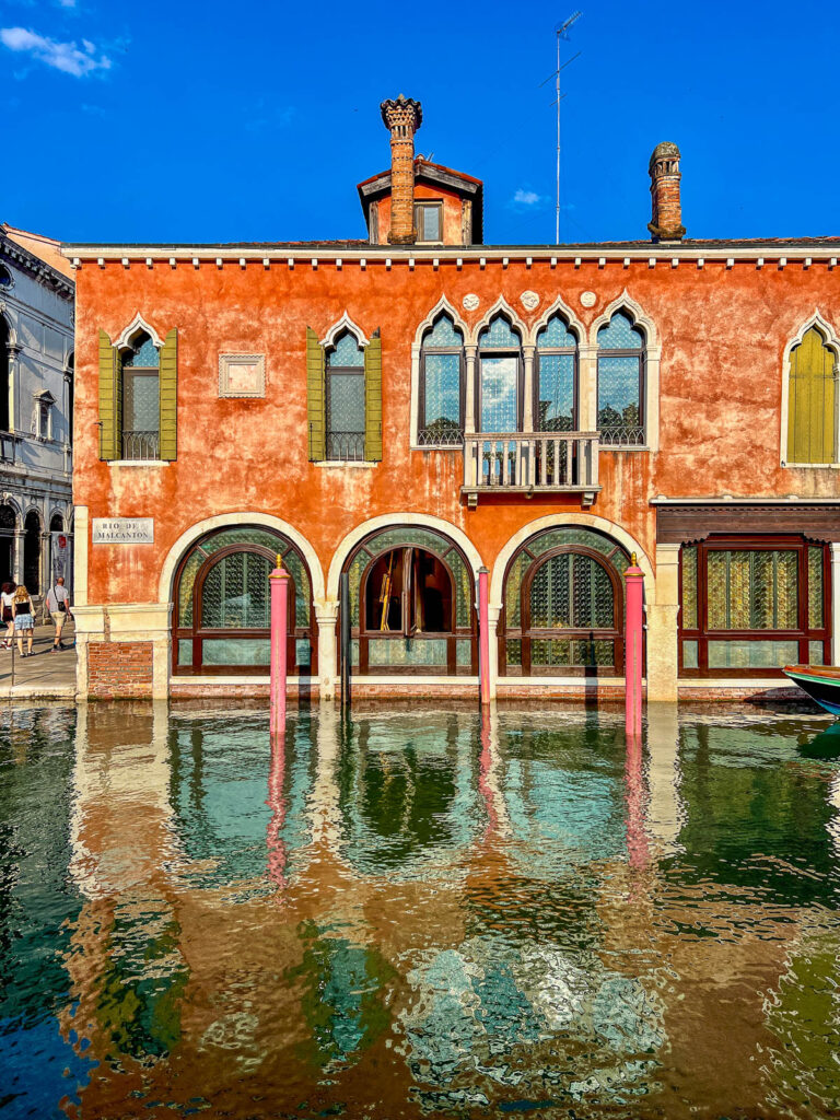 A red brick palazzo with white arched windows reflected in a Venice canal.